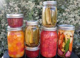 canned-food-in-jars.jpeg seven mason jars with tomatoes in one, pickle spears in two, red jam in two, a variety of vegetables in one jar with corn and other vegetables, and fruits in one jar.