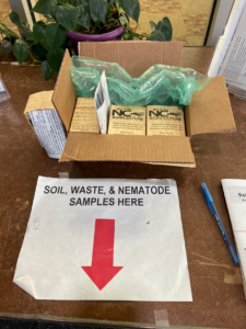 Soil , waste & nematode sample here sign on table with red arrow pointing to drop off location. 4 soil sample boxes in a box.