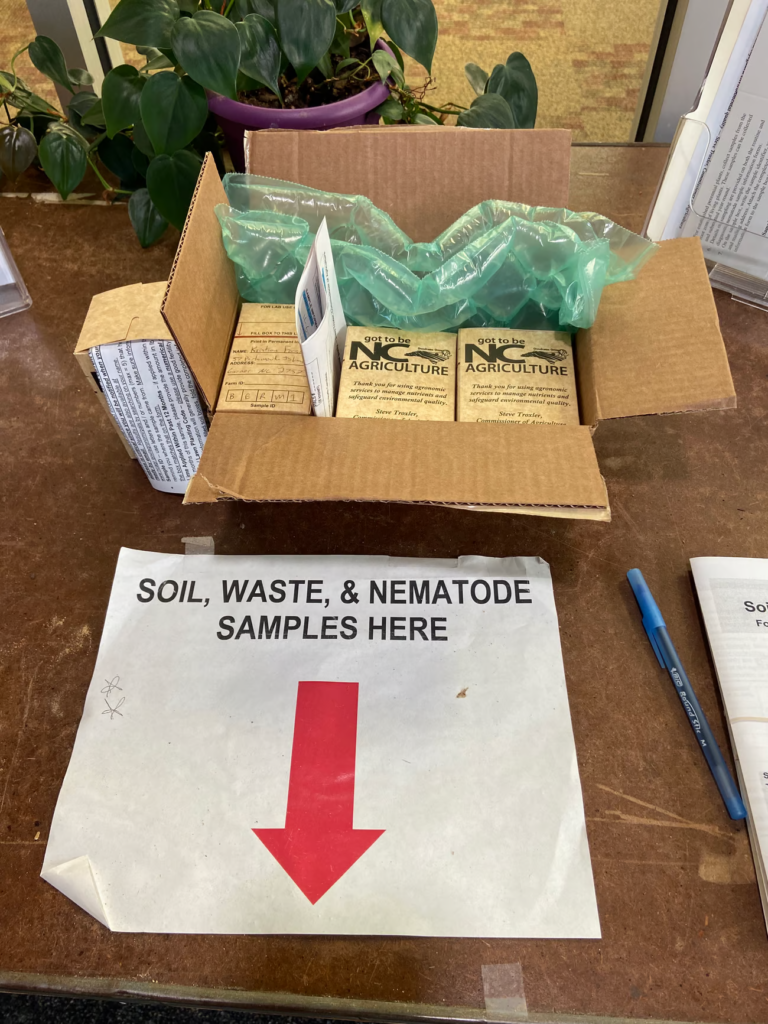 Soil , waste & nematode sample here sign on table with red arrow pointing to drop off location. 4 soil sample boxes in a box. 